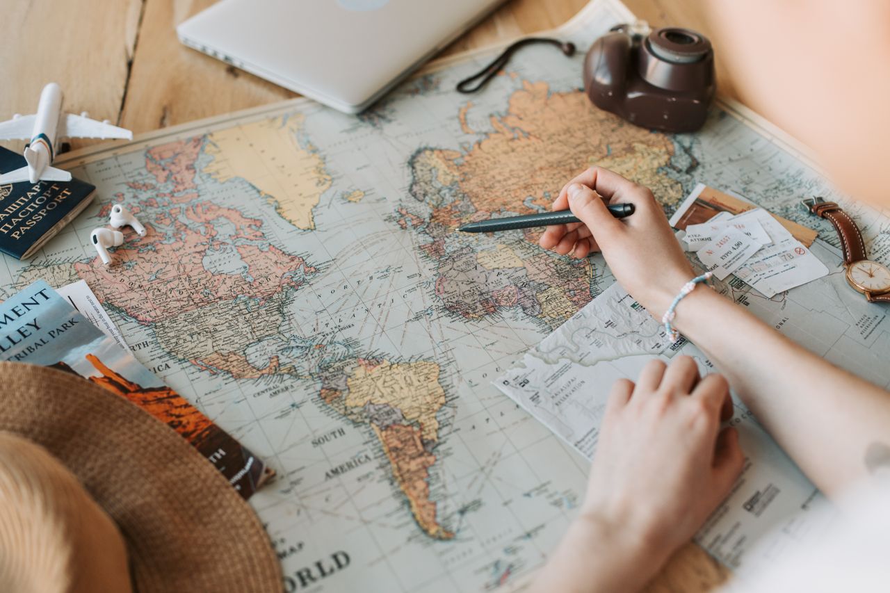 creative-03 Hands marking destinations on a world map surrounded by travel essentials like passport and camera.